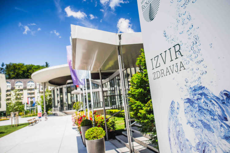 Moderna zgradba s svetlo arhitekturo in širokimi steklenimi površinami. Pred vhodom se nahajajo pisan cvetlični okras in znak