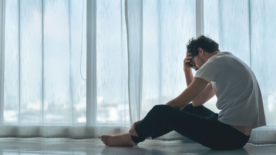 Moški sedi na tleh ob velikem oknu, ki je zavesno zakrito. Drži se za glavo in izgleda utrujen ali obremenjen.