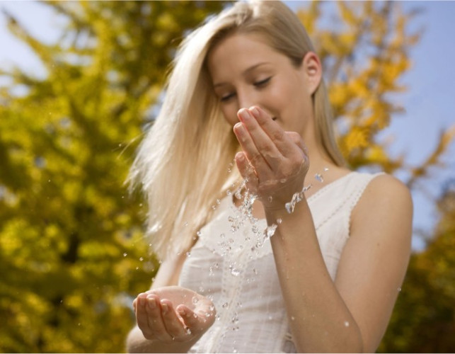 Ženska z dolgimi svetlimi lasmi stoji na prostem in z veseljem gleda v roke, iz katerih kaplja voda. V ozadju so drevesa z rumenim listjem, kar ustvarja jesenski ambient.