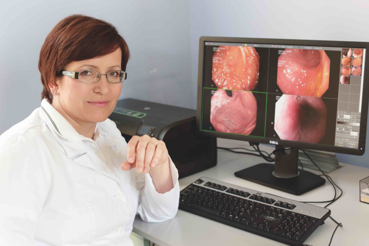 Zdravnica s kratkimi lasmi in očali sedi za računalnikom, kjer se na zaslonu prikazujejo medicinski posnetki notranjih organov. Njena roka je naslonjena na mizo, medtem ko gleda v kamero s samozavestnim nasmehom.