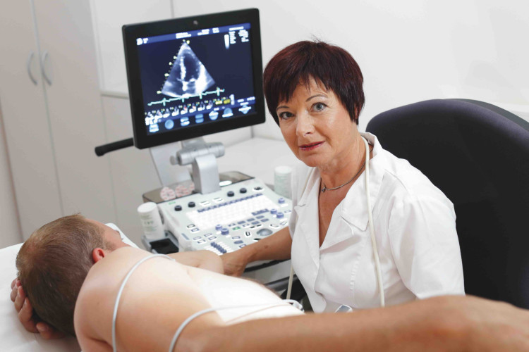 Zdravnica sedi ob ultrazvočnem aparatu in gleda v kamero. Na zaslonu je prikaz ultrazvoka, medtem ko pacient leži obrnjen na bok.