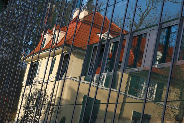 V stekleni fasadi modernega stavbe se zrcali stara hiša z rdečo streho in zelenimi okni.