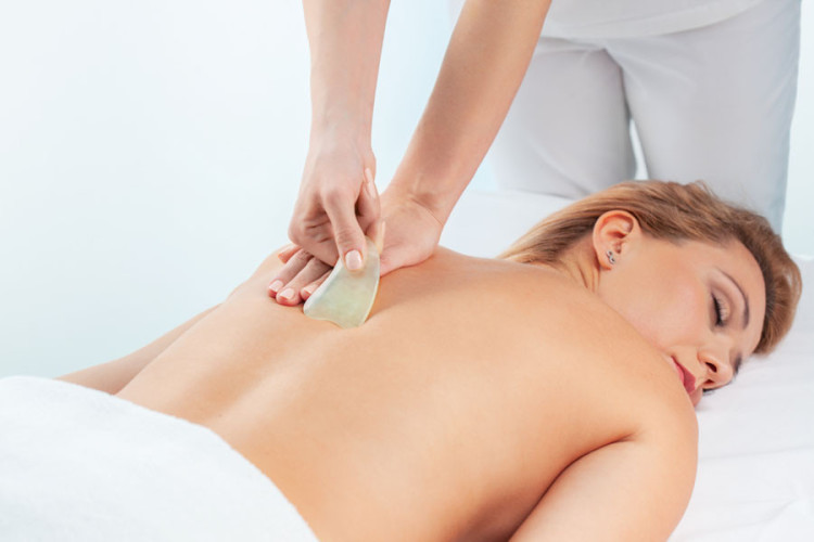 Na hrbtu ženske se izvaja masaža z uporabo kamna, medtem ko leži na masažni mizi. Ima sproščen izraz obraza in je oblečena le v brisačo.