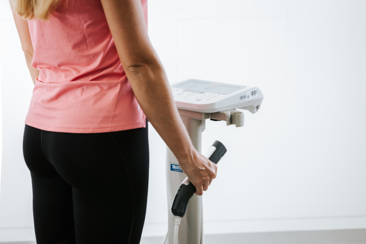Ženska v rožnati športni majici in črnih pajkicah stoji ob napravi za analizo telesne sestave in drži enega izmed ročajev.