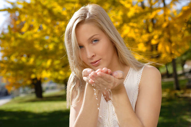 Ženska s svetlim dolgimi lasmi stoji na zelenem travniku, medtem ko z obema rokama drži nekaj kapljic vode. V ozadju so drevesa z rumenim listjem, kar ustvarja prijetno jesensko vzdušje.