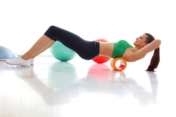 Ženska v športnih oblačilih izvaja raztezanje na vadbeni valju, medtem ko leži na trdni površini. V ozadju so fitnes žoge v različnih barvah.