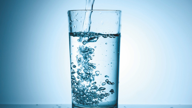 V kozarec se vliva prozorna voda, ki ustvarja mehurčke, ob tem pa odseva na mokri površini. Ozadje je osvetljeno v modrih tonih, kar daje sliki miren in svež občutek.