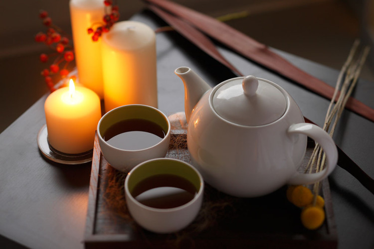 Na mizi je postavljen set za čaj, ki vključuje belo čajnico in dve skodelici s toplim čajem. Ob njem gorijo sveče, ki ustvarjajo prijeten ambient, medtem ko so dodani še suhi dekorativni elementi.