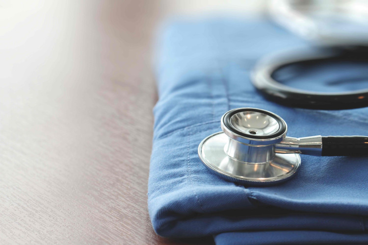 Črni stetoskop leži na modrem oblačilu na leseni površini. V ozadju so nejasne oblike, ki spominjajo na medicinske pripomočke.