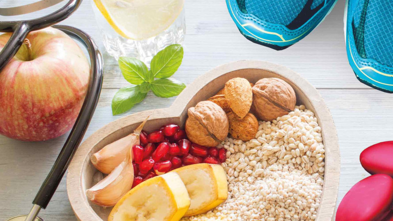 Na leseni deski v obliki srca so različne zdrave sestavine, kot so ovseni kosmiči, granatno jabolko, mandlji, lešniki in česnovi stroki. Ob strani je rdeča jabolka, kozarec limonine vode, stetoskop in modri športni čevlji.